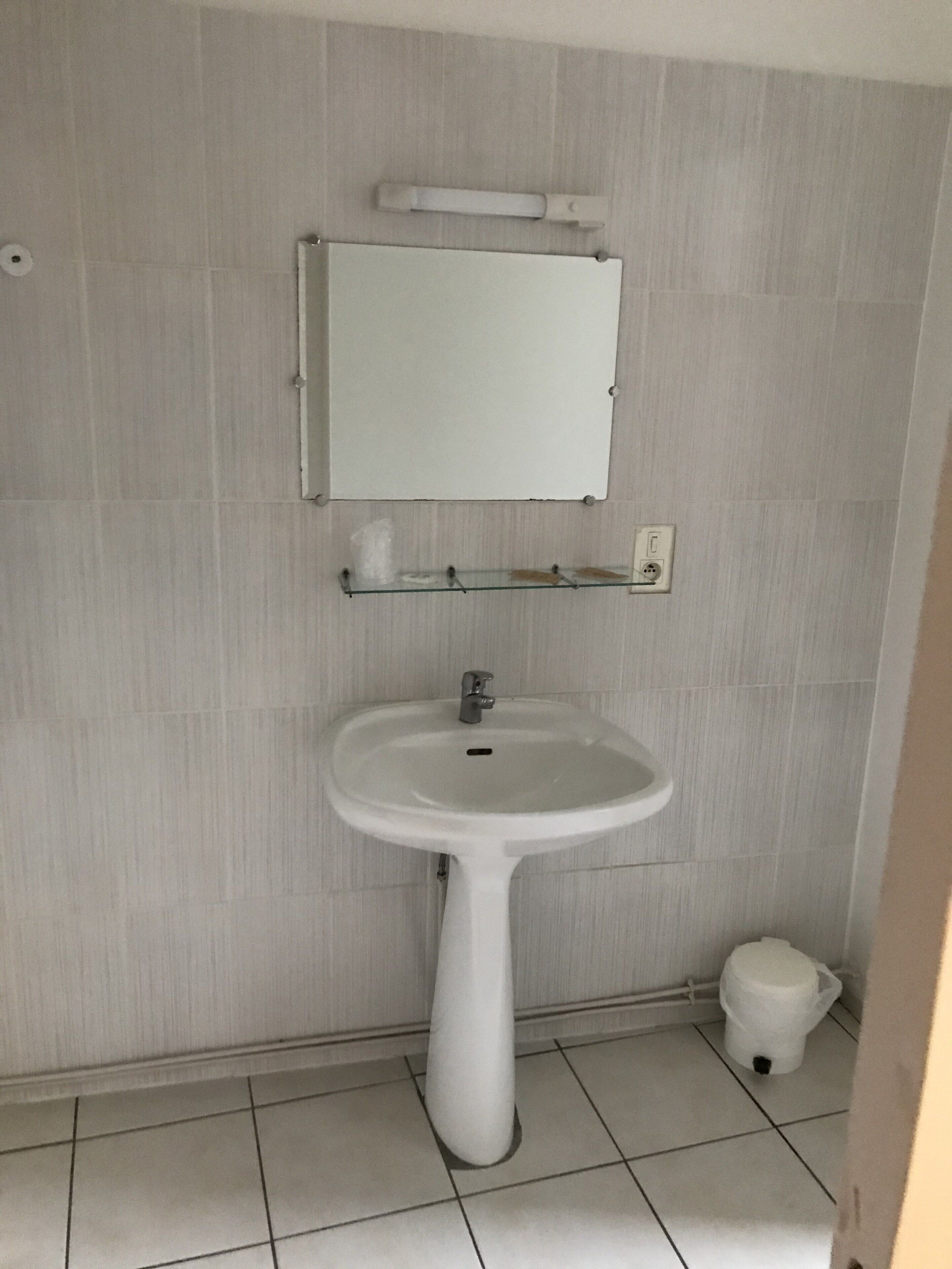 Een badkamer met een wit wastafeltje, een spiegel erboven en een klein plankje met een beker.