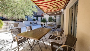 An outdoor patio with tables and chairs under a striped awning, a pool, and a building with a door and windows. - Hotel de la Gare – urban by balladins (Chateauroux)