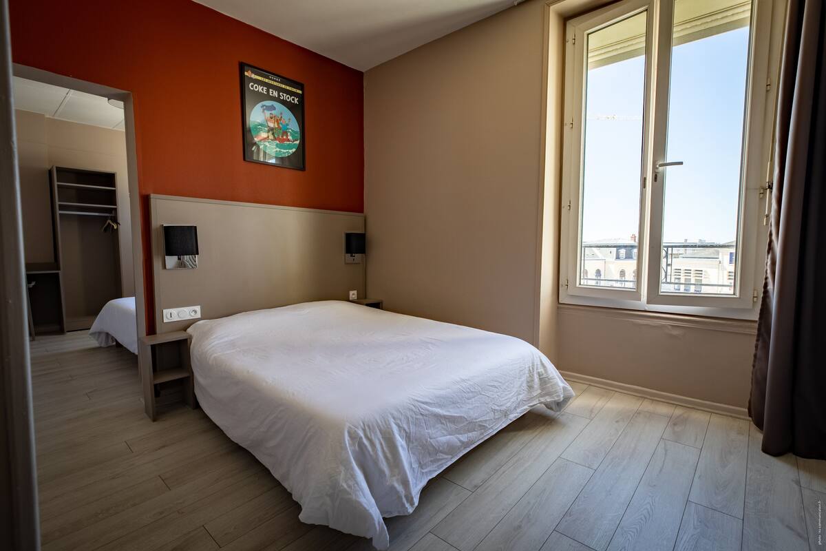 A hotel room with a bed, a nightstand, a wall-mounted TV, a window with curtains, and a poster on the wall.