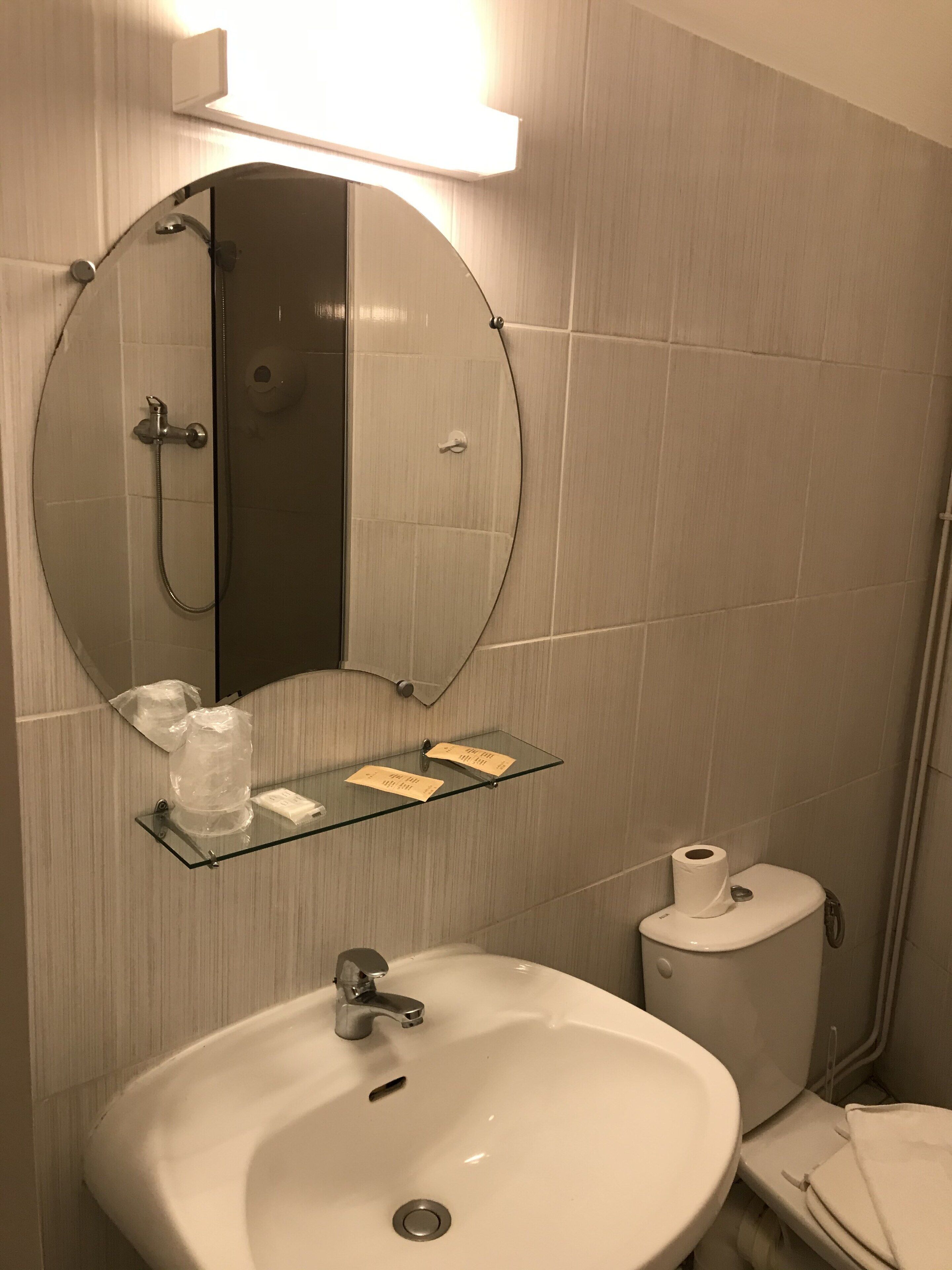 Une salle de bain avec un miroir rond, un lavabo, des toilettes et une étagère en verre où sont rangés les articles de toilette.