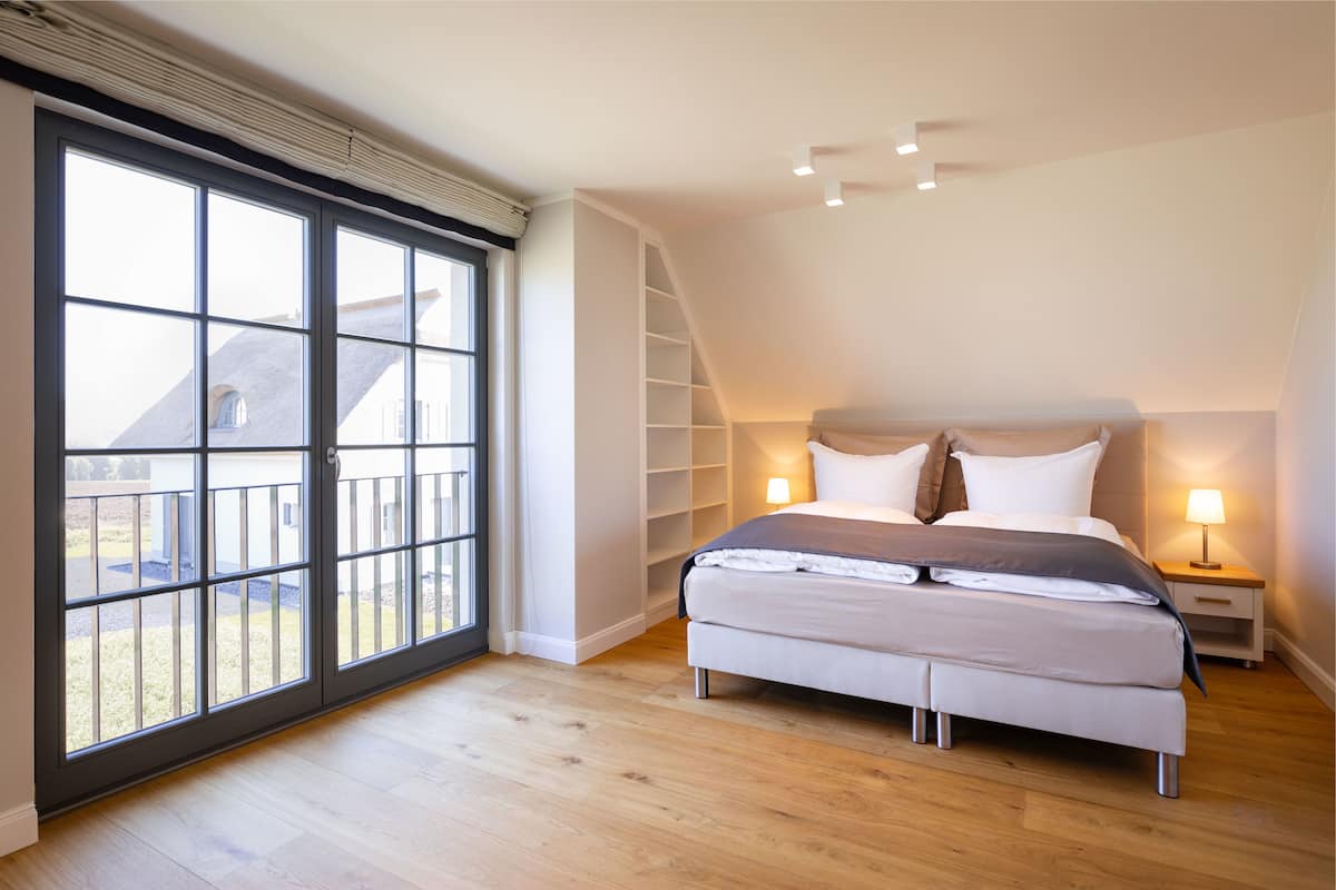 A bedroom with a large window, a bed, a nightstand, and a built-in shelving unit.