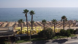 Une plage bordée de rangées de parasols et de palmiers, une route au premier plan et la mer en arrière-plan.