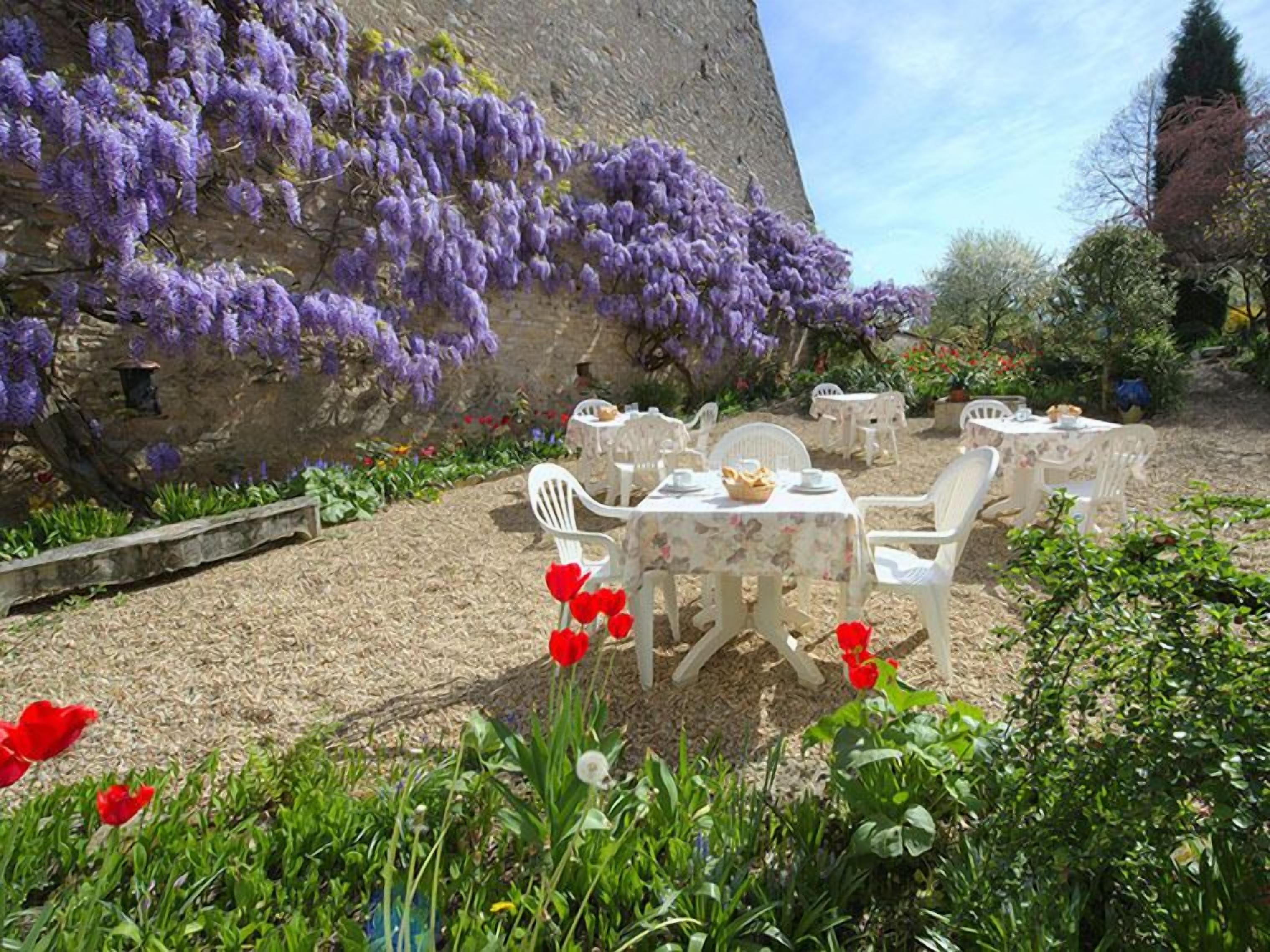 Un espace extérieur aménagé avec des chaises et des tables en plastique blanc, entouré d’un jardin où poussent des tulipes rouges et des fleurs violettes.