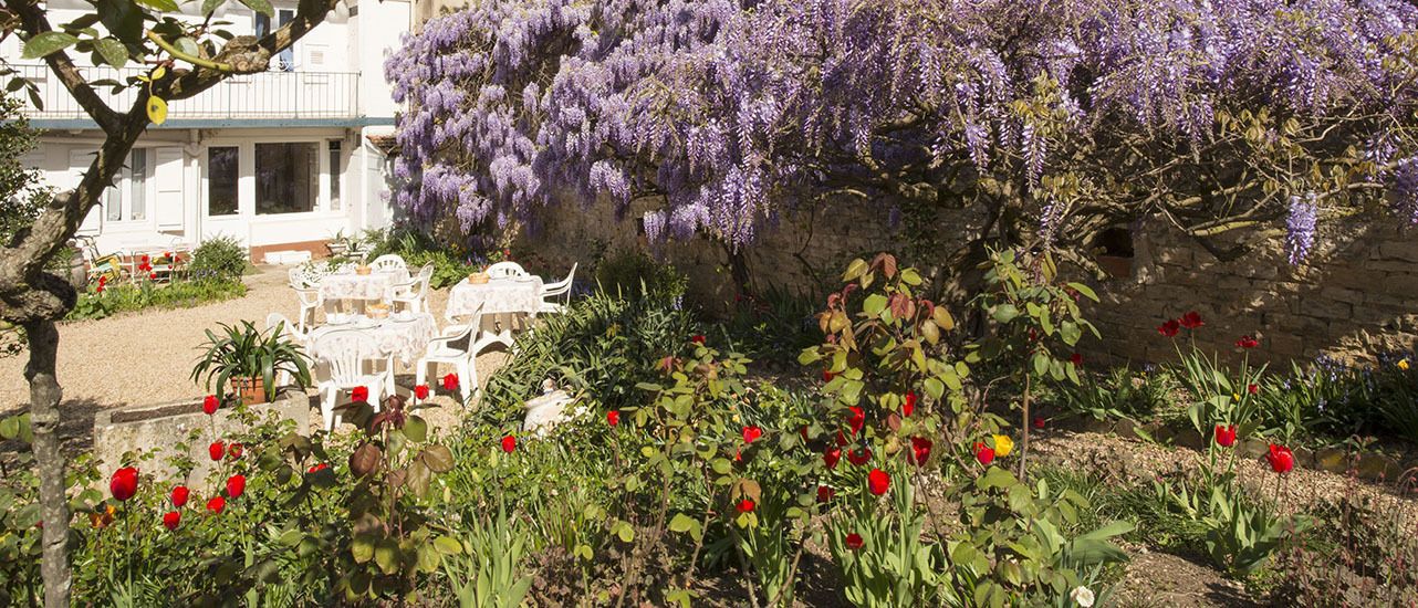 A garden with white outdoor furniture, a variety of flowers, and purple wisteria vines.