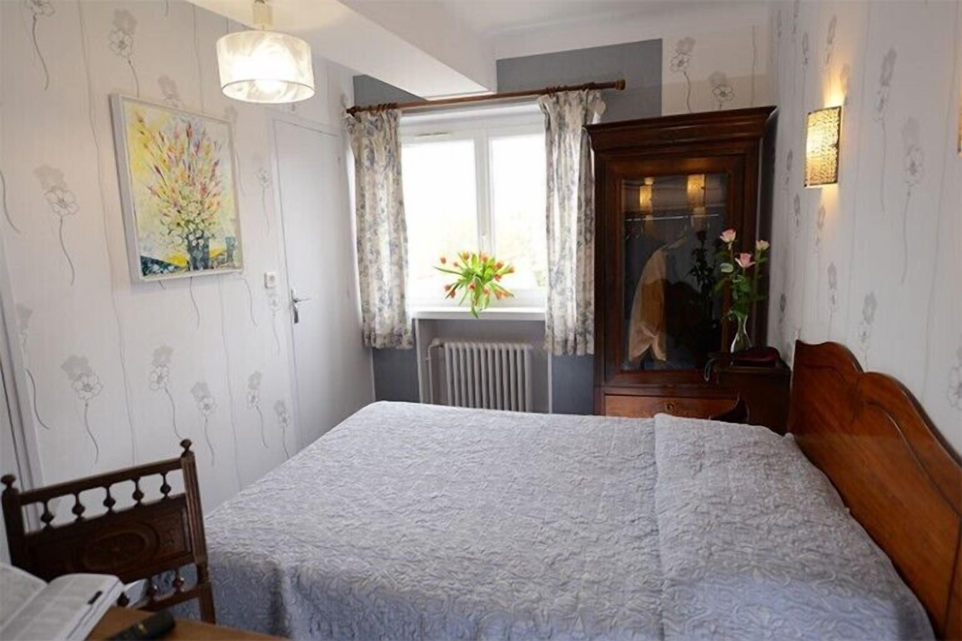 A bedroom with a bed, a wooden headboard, a window with curtains, a radiator, a wall-mounted light, and a painting.