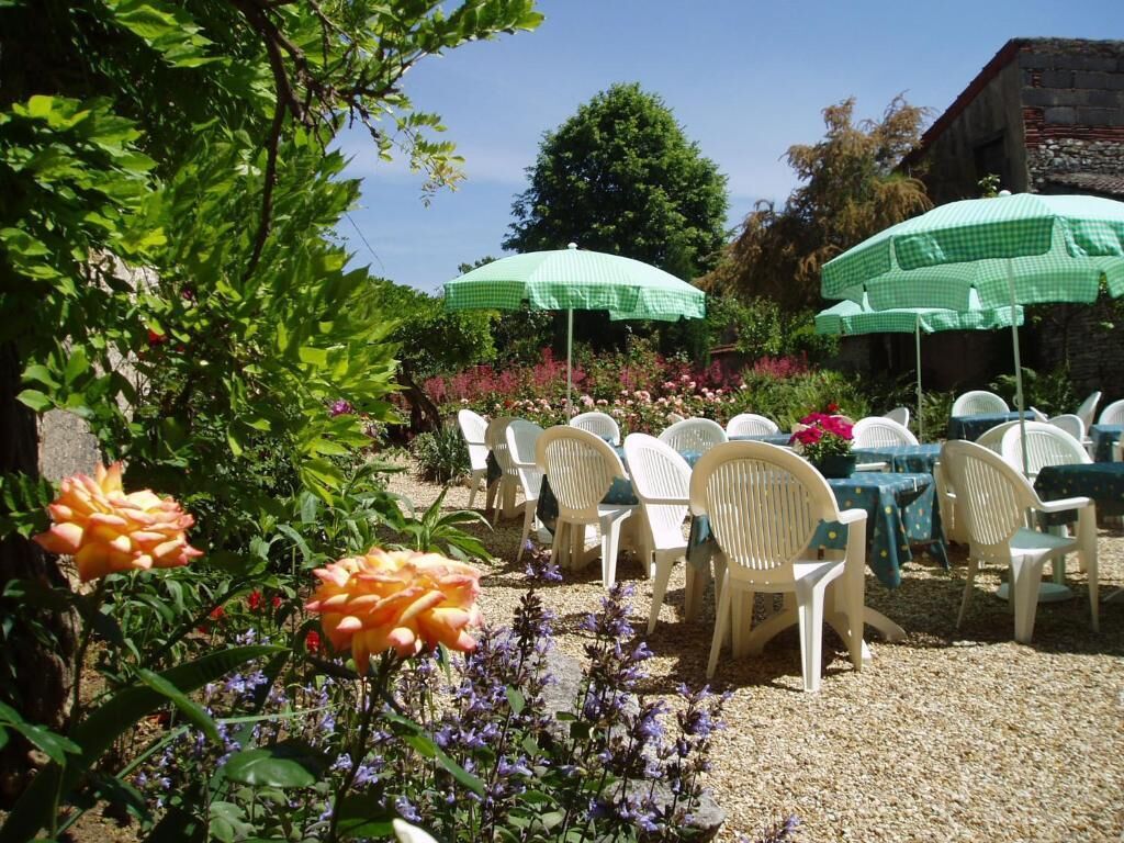 Ein Sitzbereich im Freien mit weißen Plastikstühlen und -tischen, grün-karierten Sonnenschirmen sowie einem Garten mit Rosen und violetten Blumen.