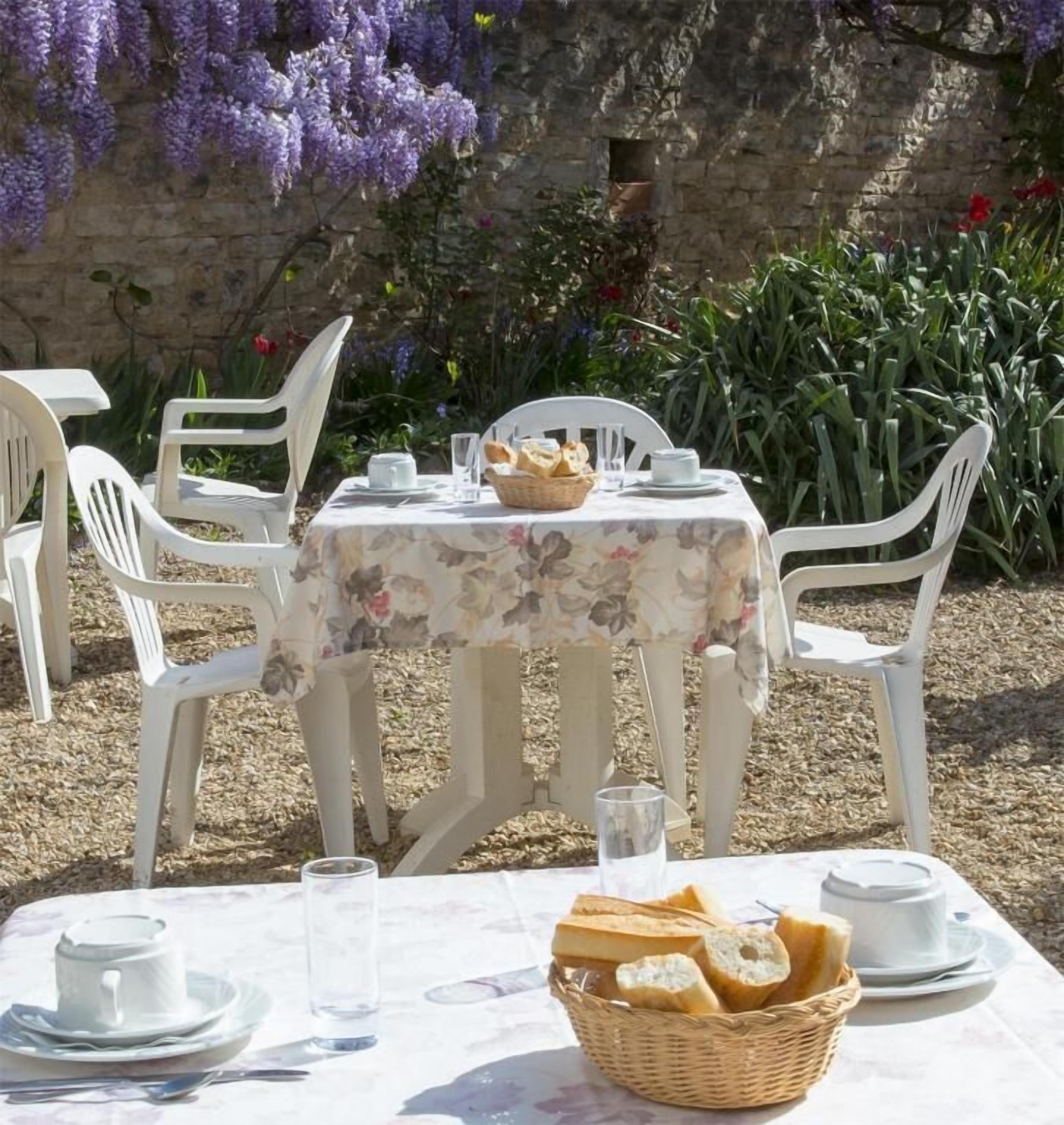 An outdoor dining area with a table set for two, featuring white plastic chairs, a floral tablecloth, and a basket of bread.