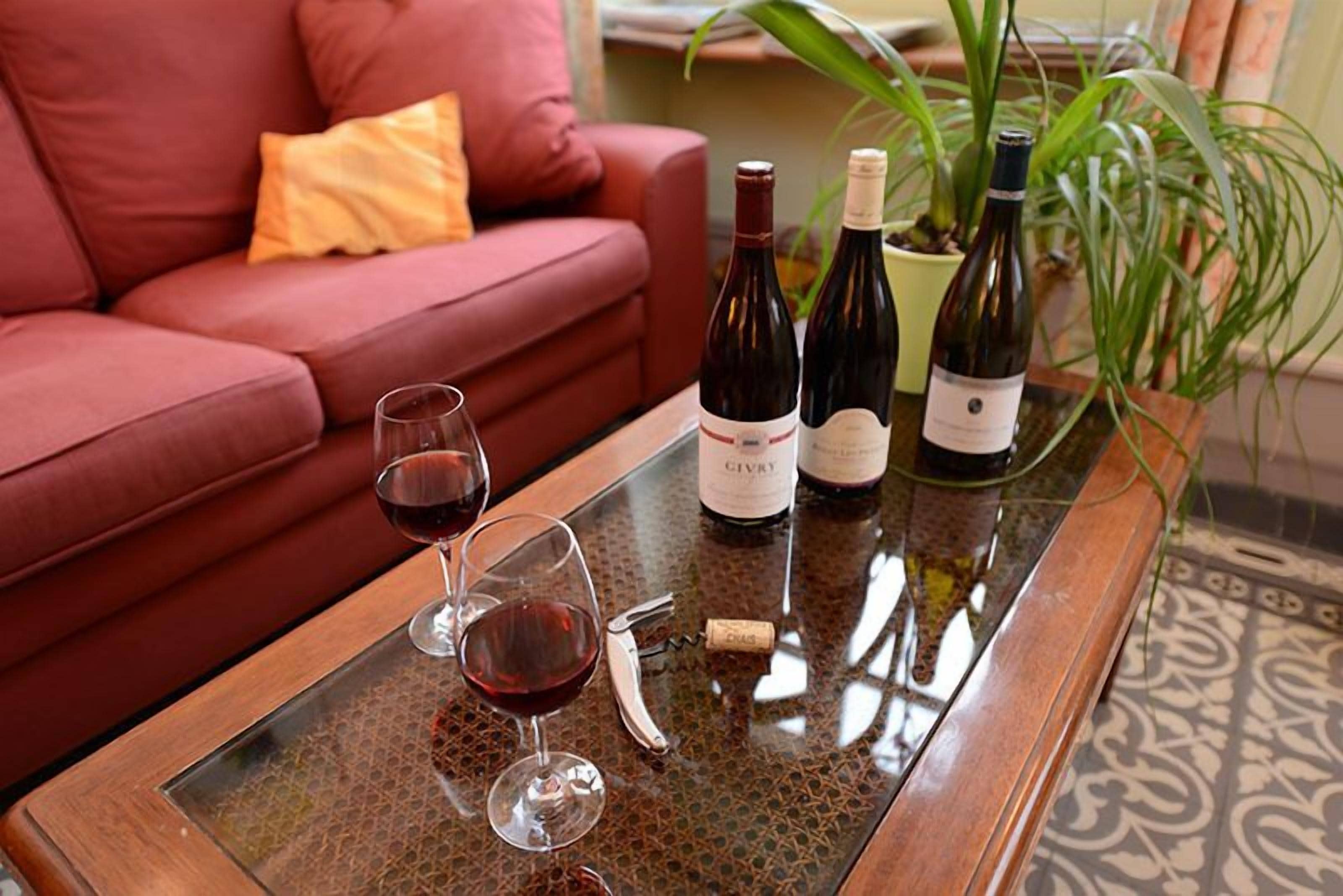 Una mesa de vidrio con botellas y copas de vino, un sacacorchos, y un sofá de cuero rojo con un cojín amarillo al fondo.
