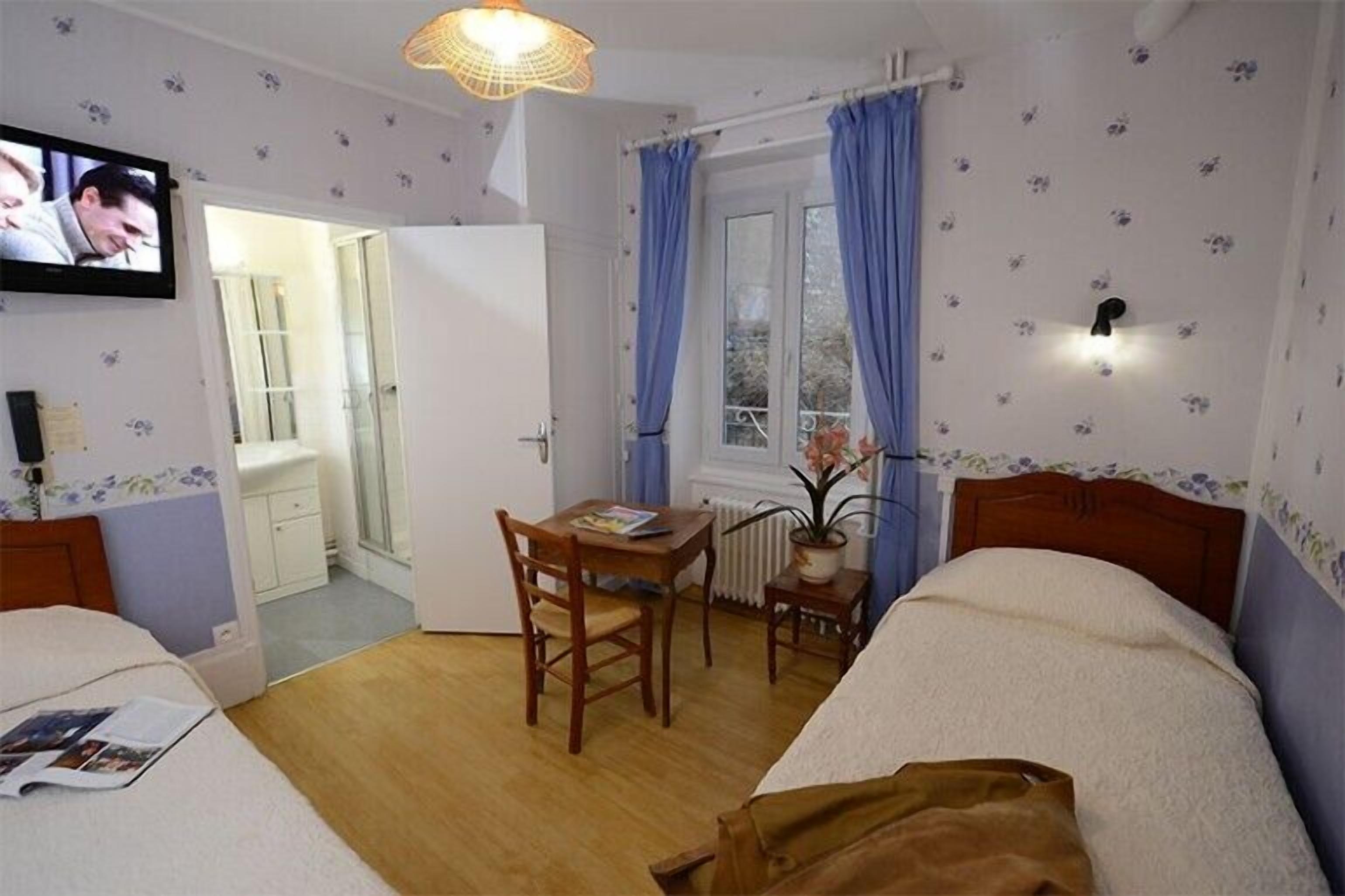 Quarto de hotel com duas camas, uma mesa de madeira, uma cadeira, uma mesinha com uma planta, um radiador e uma televisão fixada na parede.