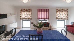 Um apartamento moderno com dois quartos e uma varanda. Possui uma sala de jantar com toalha de mesa azul, uma televisão e um vaso decorativo com flores.