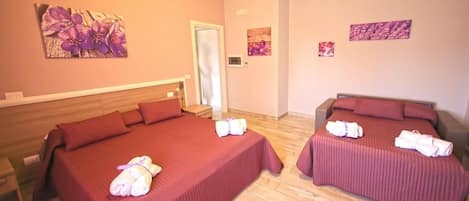 A hotel room with two beds, each with a red bedspread and towels. There are floral wall art pieces and a door in the background.
