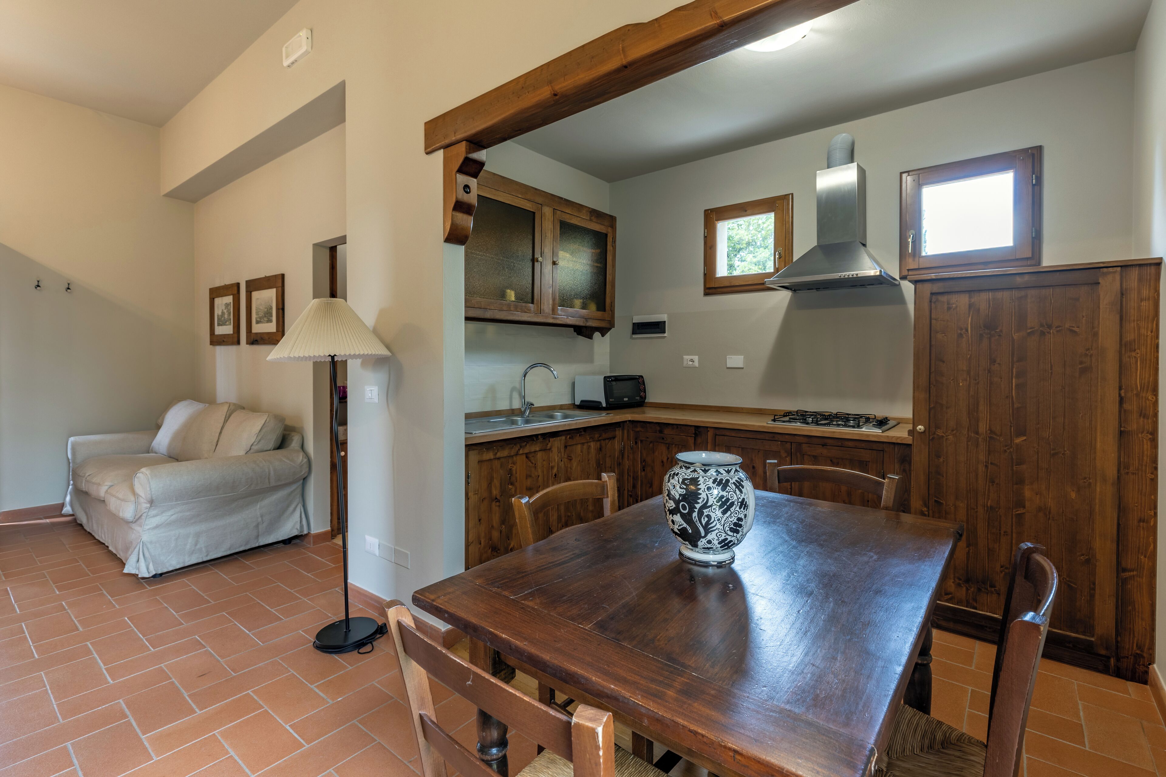 Uma sala de jantar com uma mesa de madeira, cadeiras e um vaso. Há um sofá e um abajur na sala adjacente. Cozinha visível com armários de madeira e uma janela.
