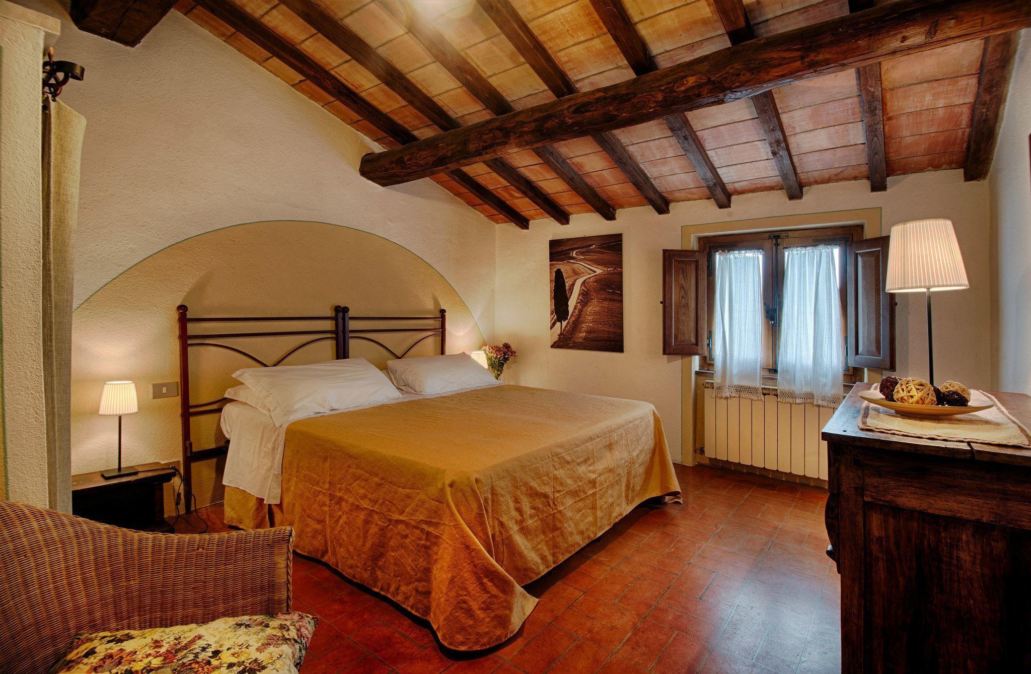 A cozy bedroom with a bed, a wooden ceiling, a window with curtains, and a bedside table with a lamp.