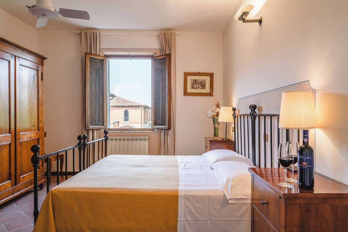 Une chambre d’hôtel avec un lit, une commode en bois, une table de chevet avec une lampe, une bouteille de vin et une fenêtre avec des rideaux.