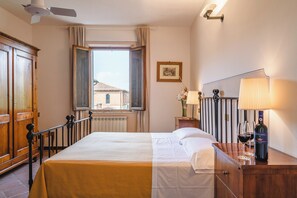 A hotel room with a bed, a wooden dresser, a nightstand with a lamp, a bottle of wine, and a window with curtains. - Agriturismo Poggio Bonelli (Castelnuovo Berardenga)