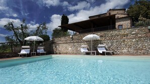 Una piscina con sillones de descanso y sombrillas, paredes de piedra y un edificio con techo inclinado.