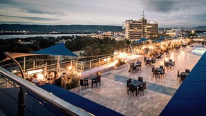Una terraza al aire libre con mesas y sillas, con vistas a un paisaje urbano y una masa de agua.