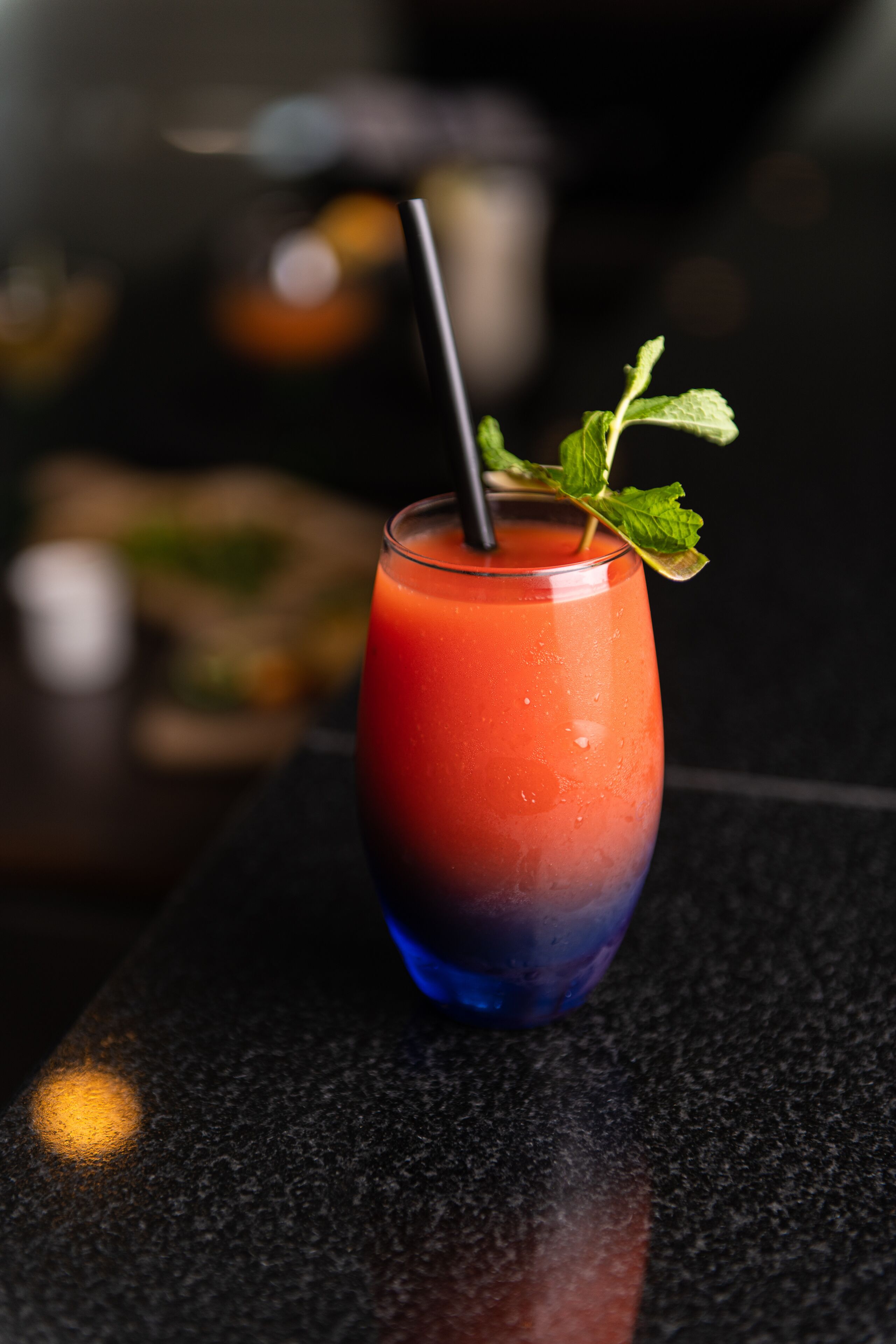 Un vaso de bebida roja y azul con pajita negra y una hoja de menta como adorno sobre una encimera oscura.