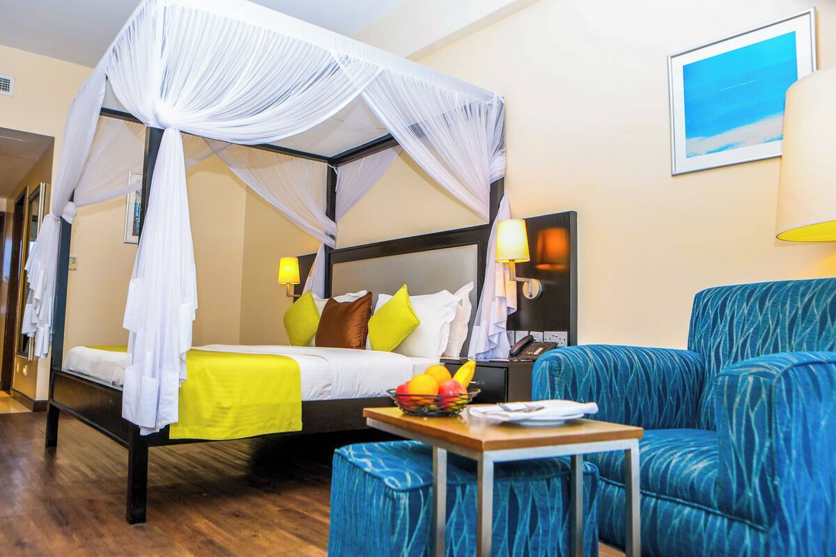 Ein Hotelzimmer mit Himmelbett, einem Nachttisch mit Obstschale und einem blauen, gepolsterten Sessel.