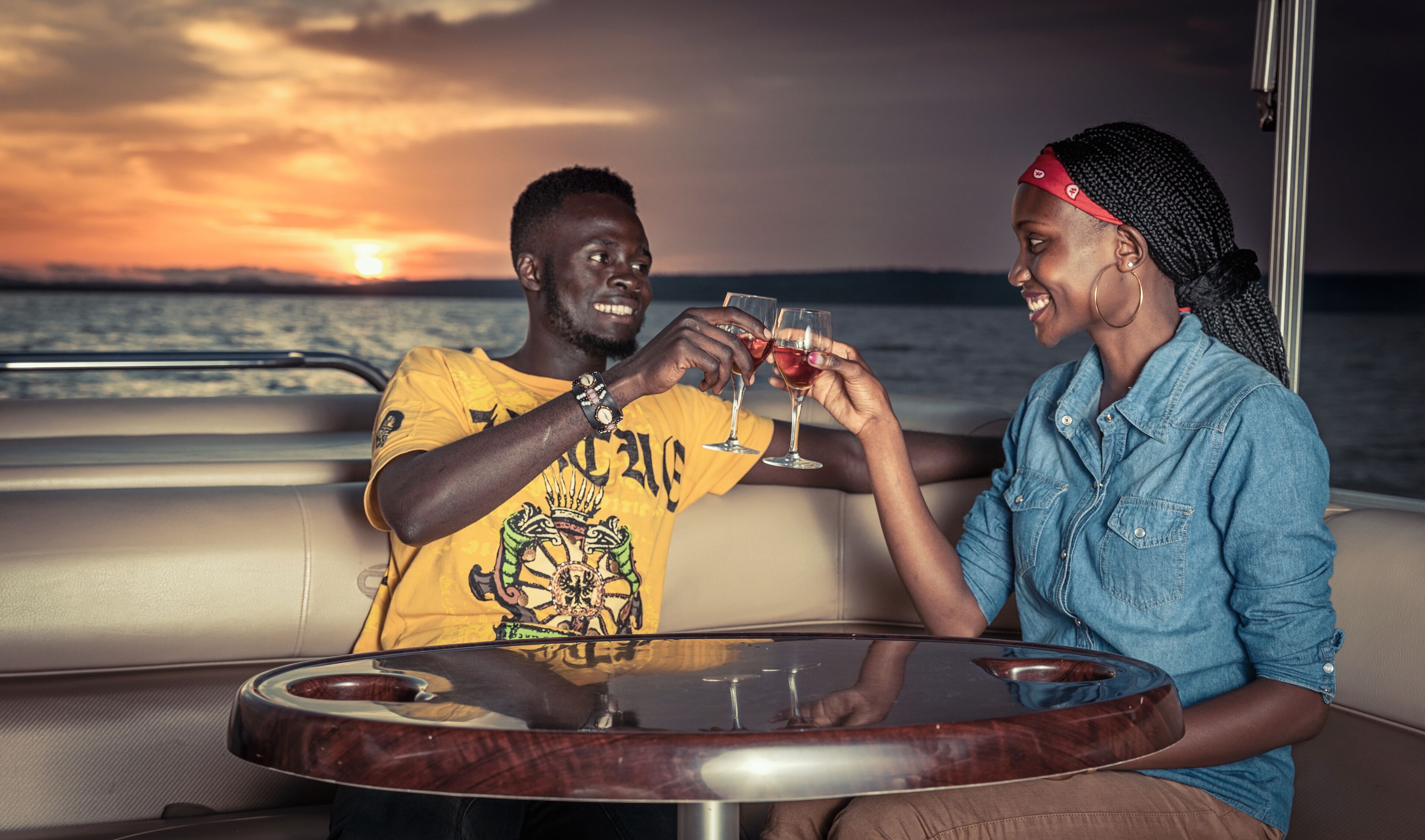 Two people toasting with drinks at a round table on a boat with a sunset view.