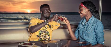 Two people toasting with drinks at a round table on a boat with a sunset view.