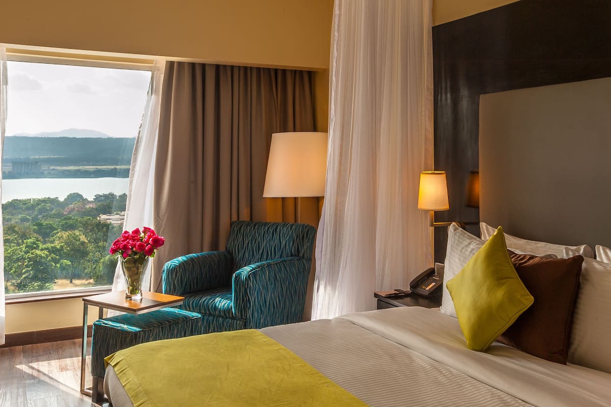 Ein Hotelzimmer mit Bett, Stuhl, Tisch mit Blumenvase, Lampe und Blick auf einen See.