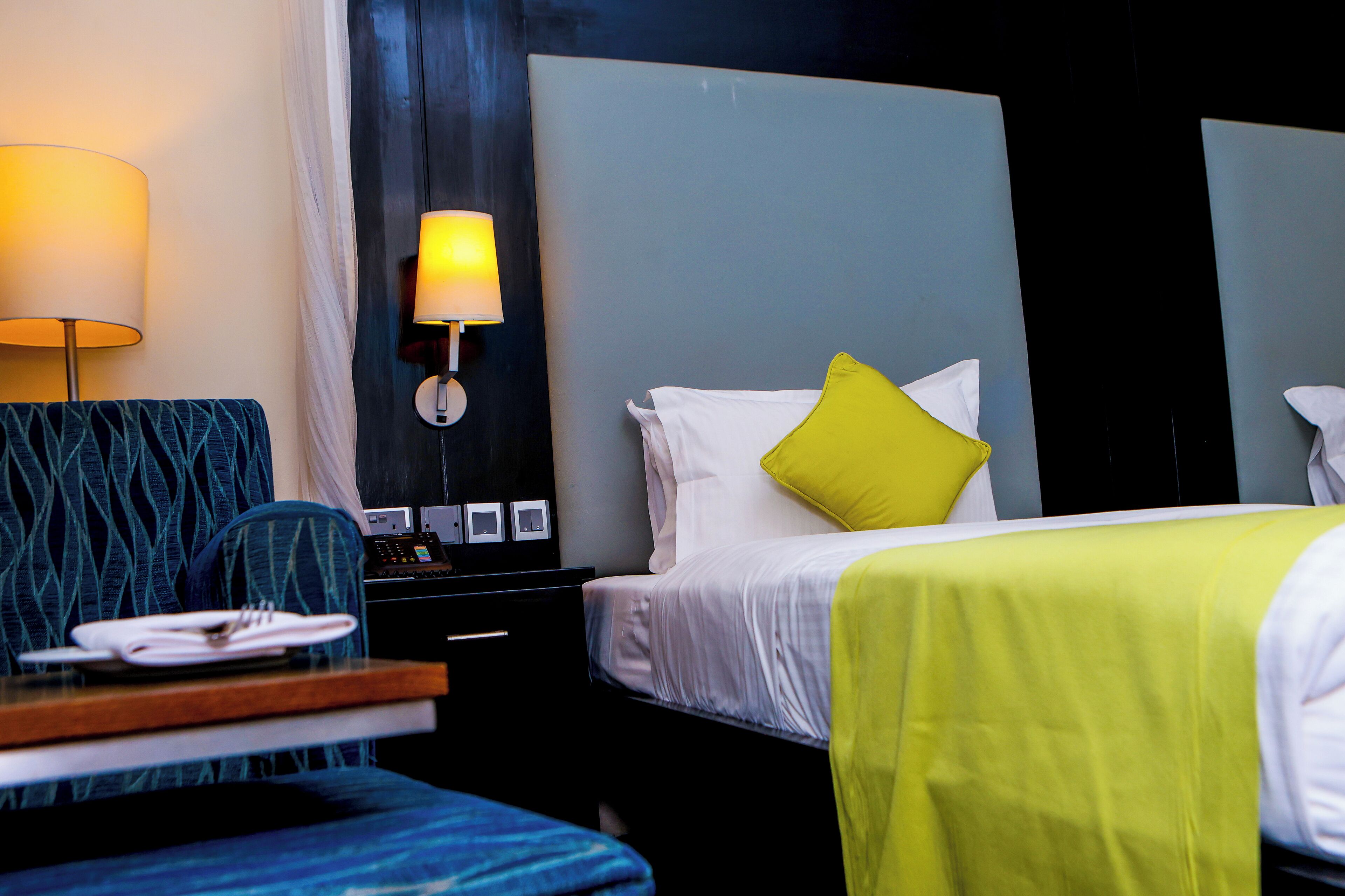 A hotel room with a bed, a bedside table with a lamp, a chair, and a desk with a plate of folded towels.