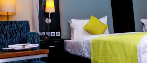 A hotel room with a bed, a bedside table with a lamp, a chair, and a desk with a plate of folded towels.