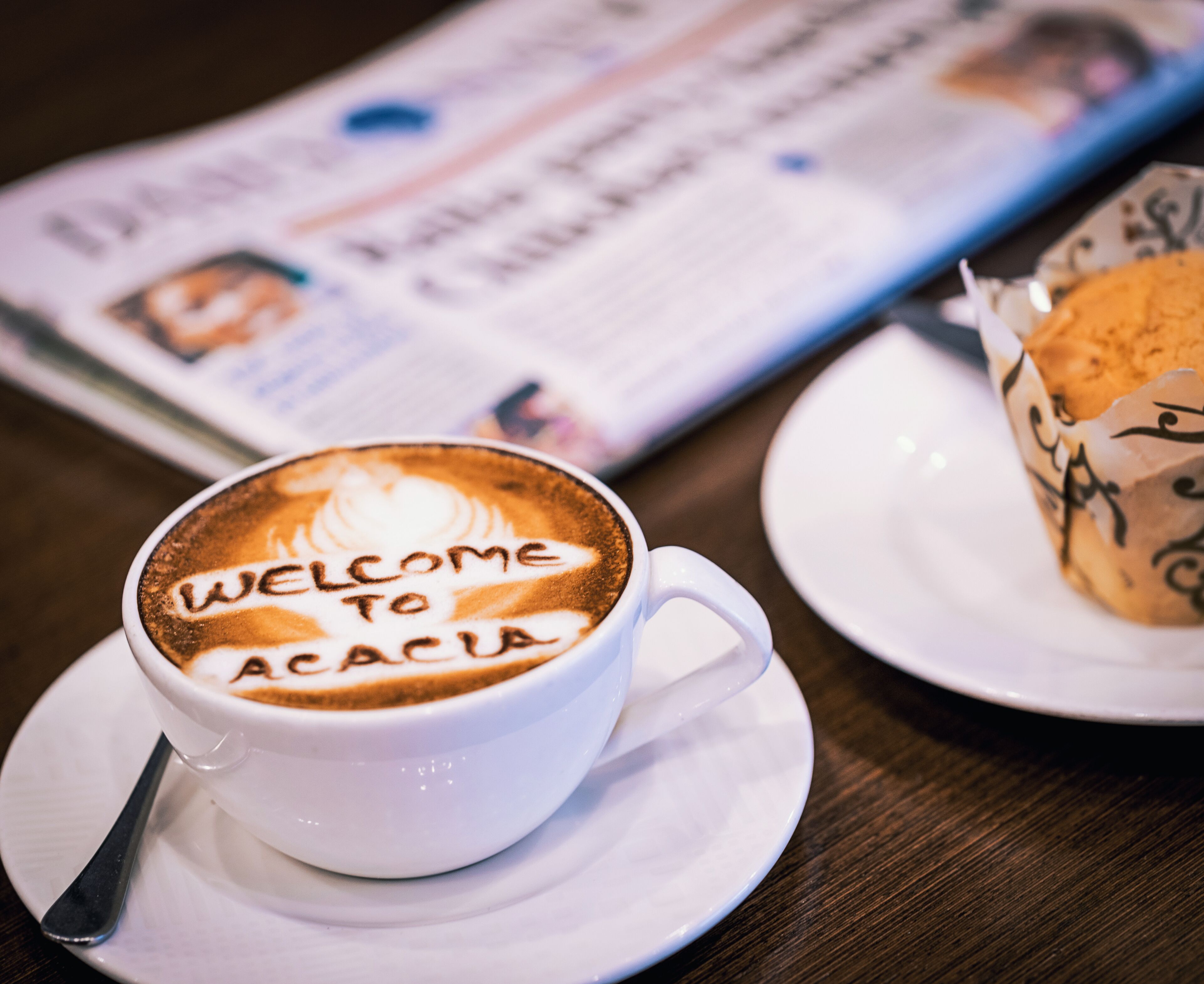 A cup of coffee with latte art reading "Welcome to Acacia" on a white saucer, a newspaper, and a plate with a pastry on a wooden table.
