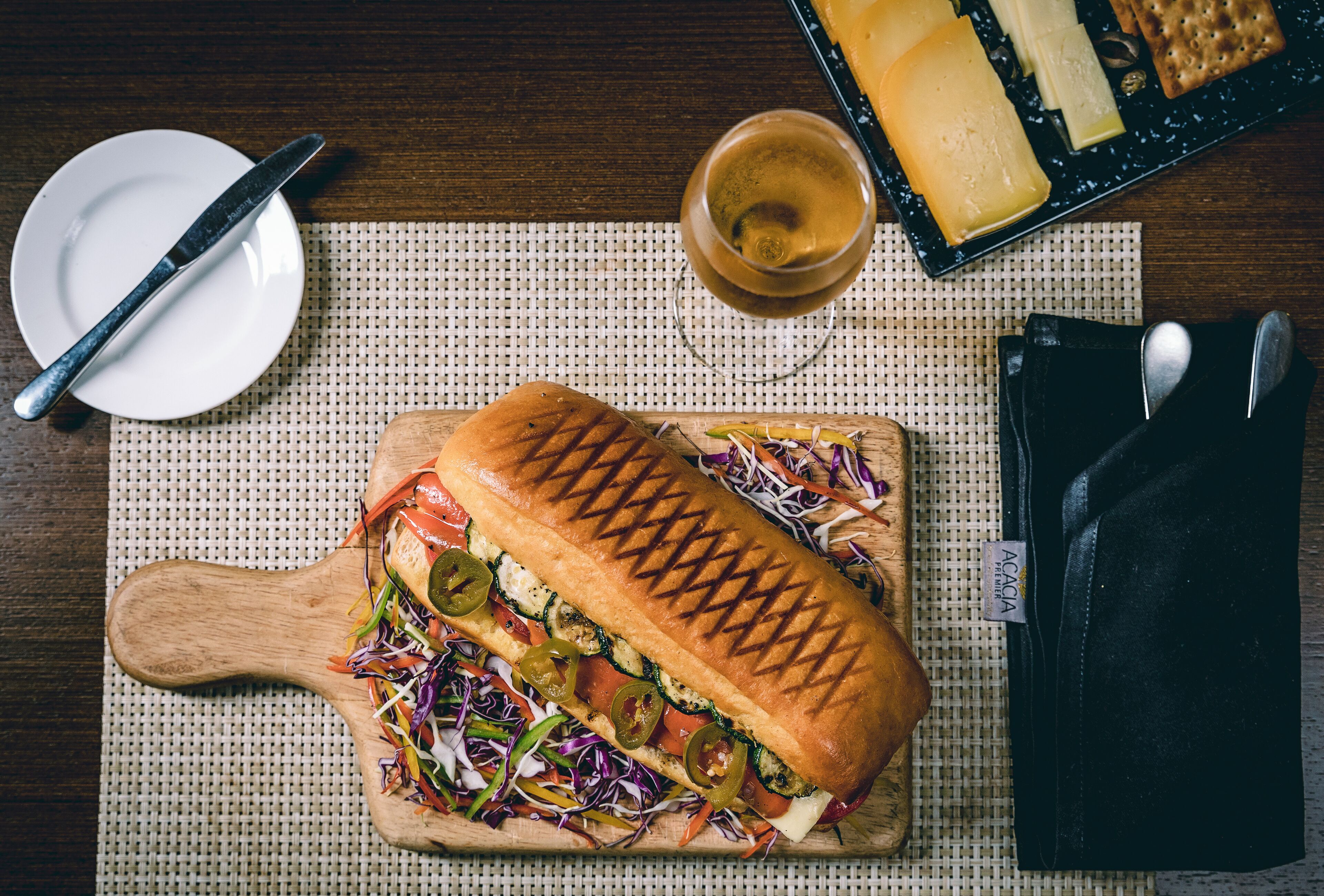 A sandwich on a wooden board, a plate with a knife, a glass of drink, and a cheese board on a table.