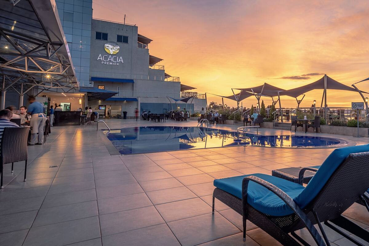 Ein Poolbereich auf dem Dach mit Liegestühlen und Sonnenschirmen, ein Gebäude mit der Beschriftung 'Academia Premier' und Blick auf den Sonnenuntergang.