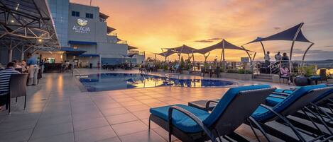 A rooftop pool area with lounge chairs and umbrellas, a building with 'Academia Premier' signage, and a sunset view.