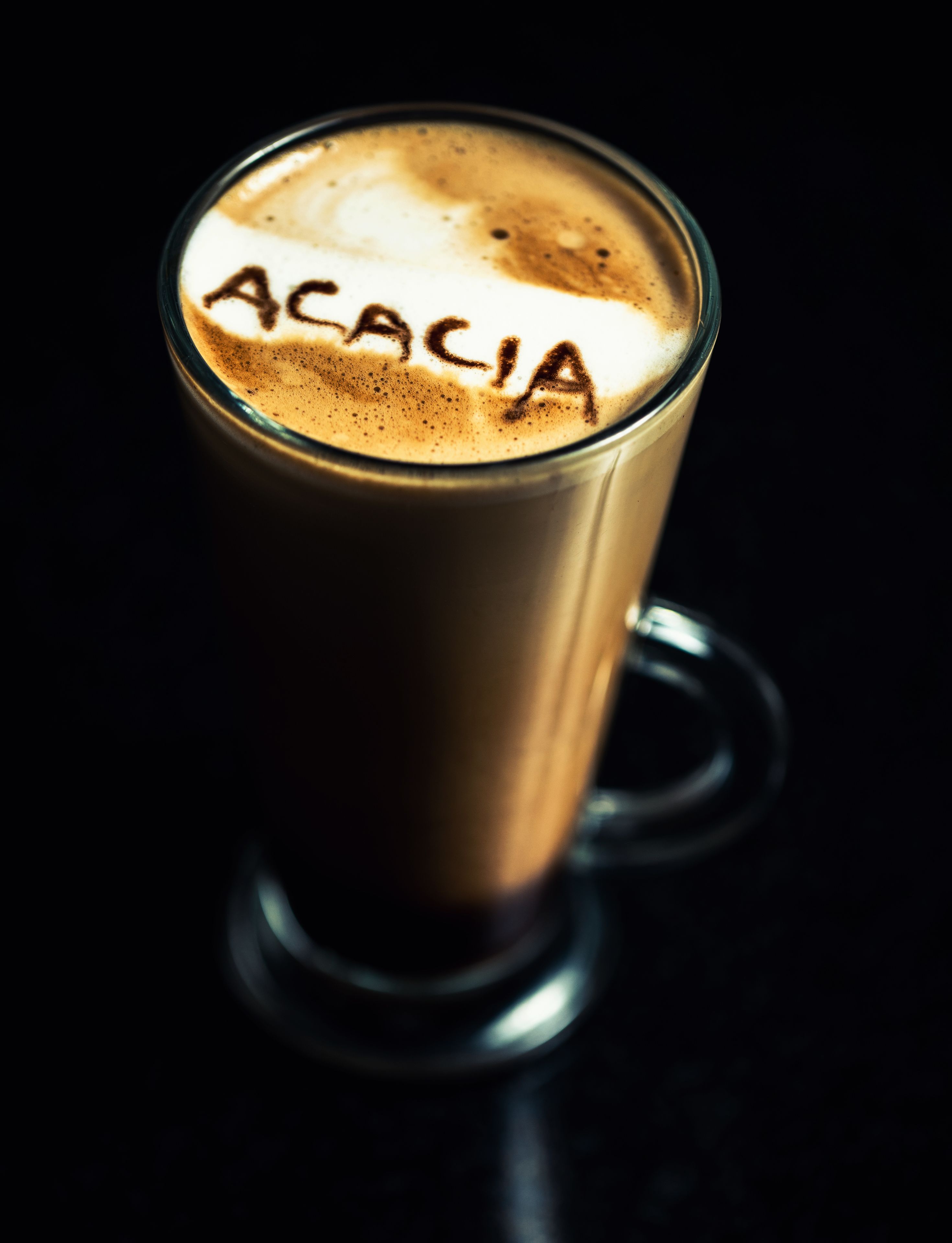 A glass cup with a dark liquid and a light-colored foam on top, with the word "ACACIA" written in foam.