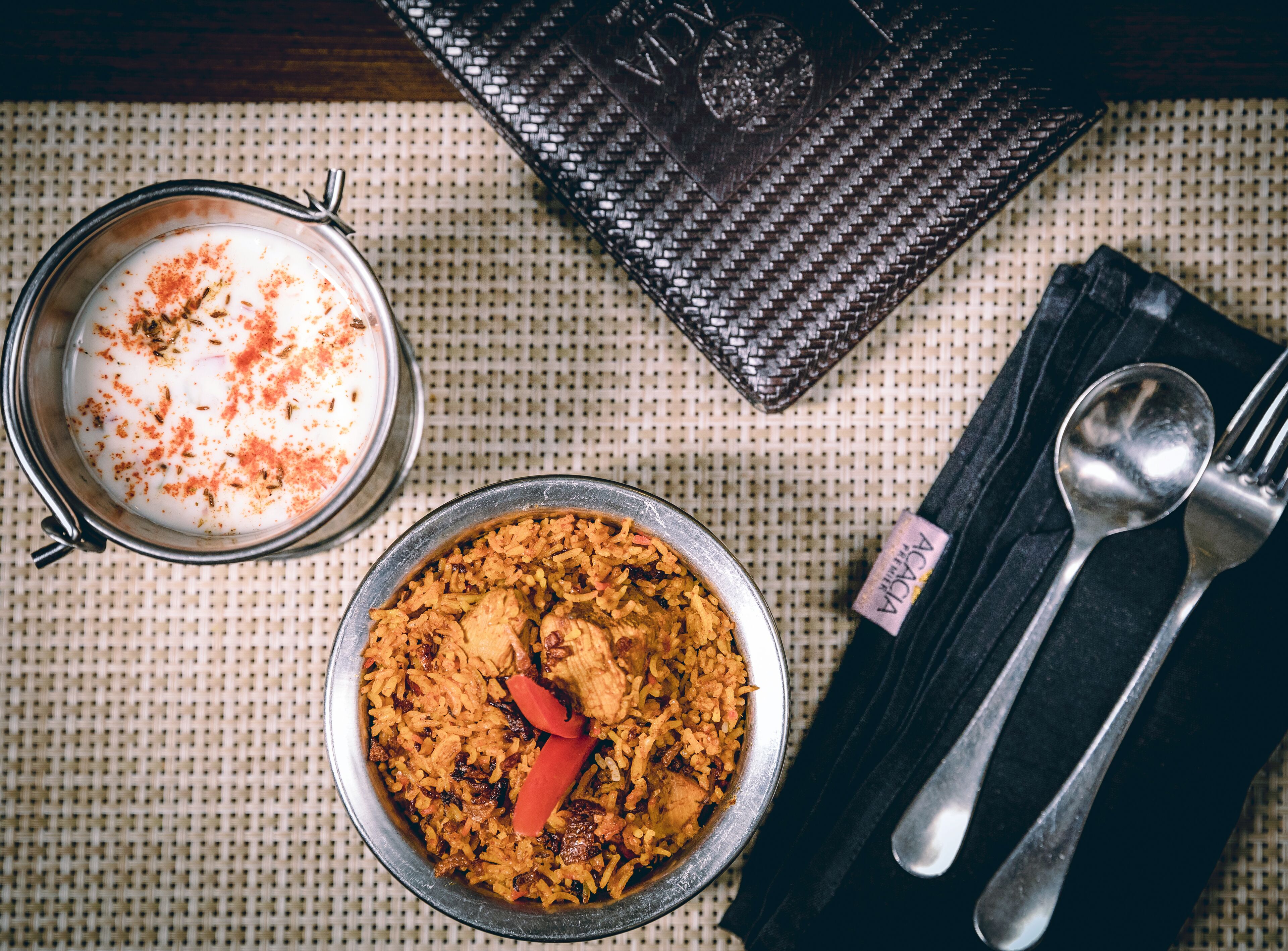 Un plato de arroz con carne, un tazón de yogur con especias, y una cuchara y un tenedor sobre un mantel negro.
