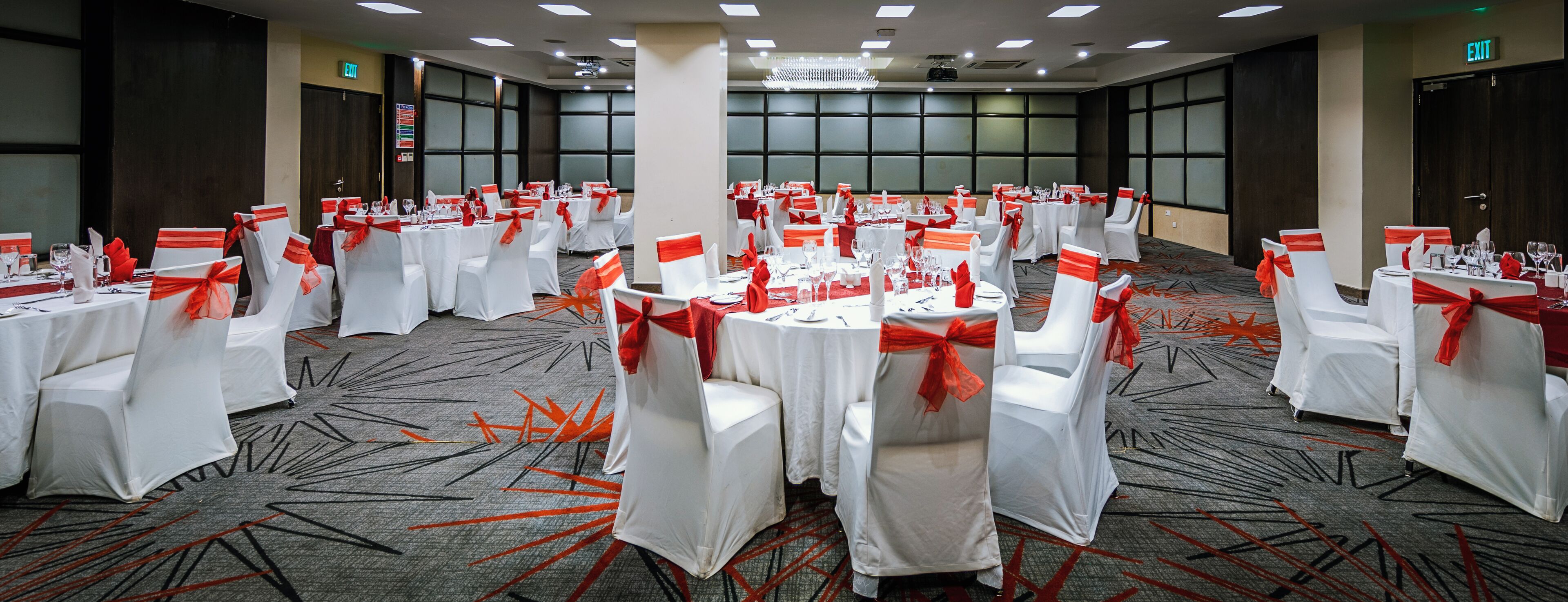 A banquet hall with round tables, white chairs with red sashes, and a carpeted floor.