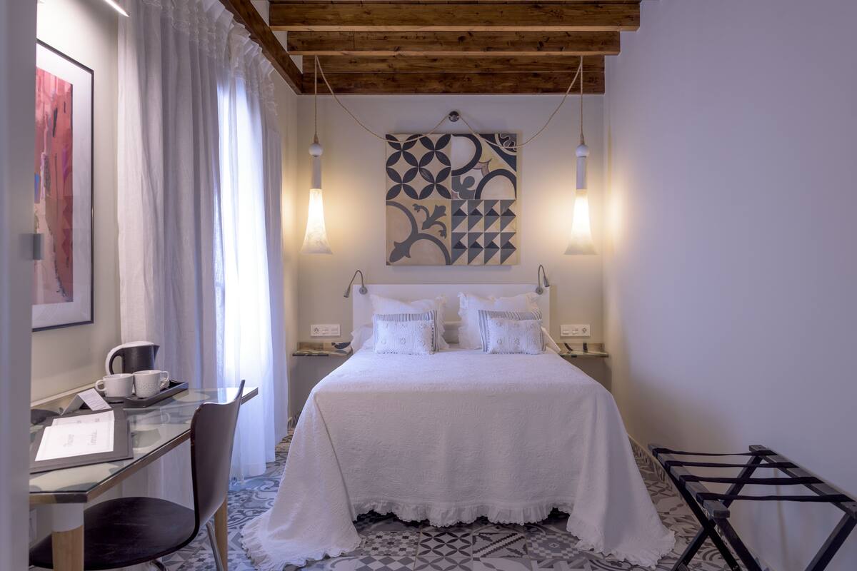 A bedroom with a bed, a desk with a chair, a hanging light fixture, and a wall art piece.