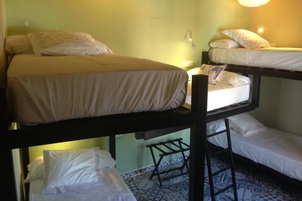 A dormitory-style room with bunk beds, a small table, and a wall-mounted lamp.