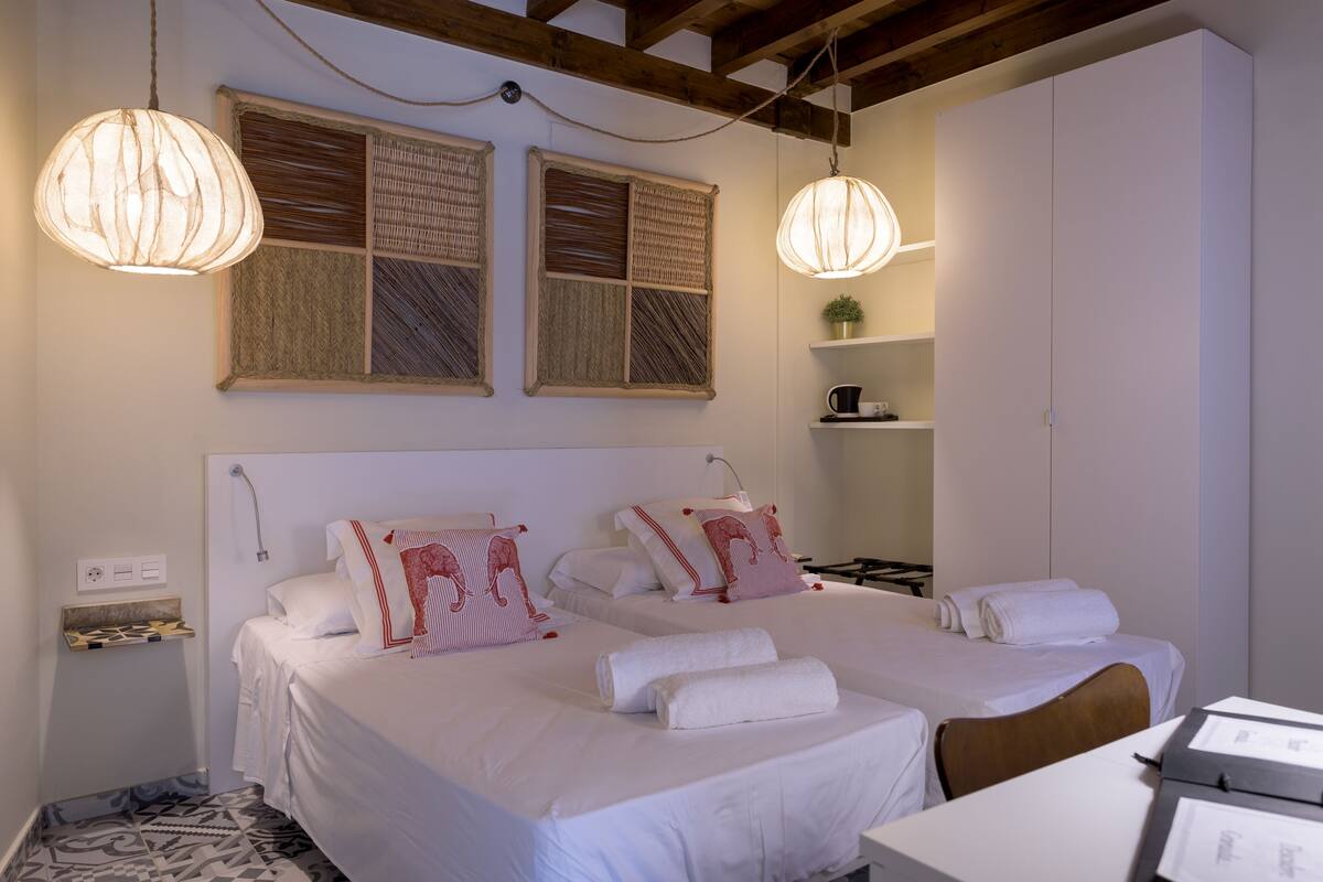 A hotel room with two beds, a small table, a shelf with a plant, and a hanging light fixture.