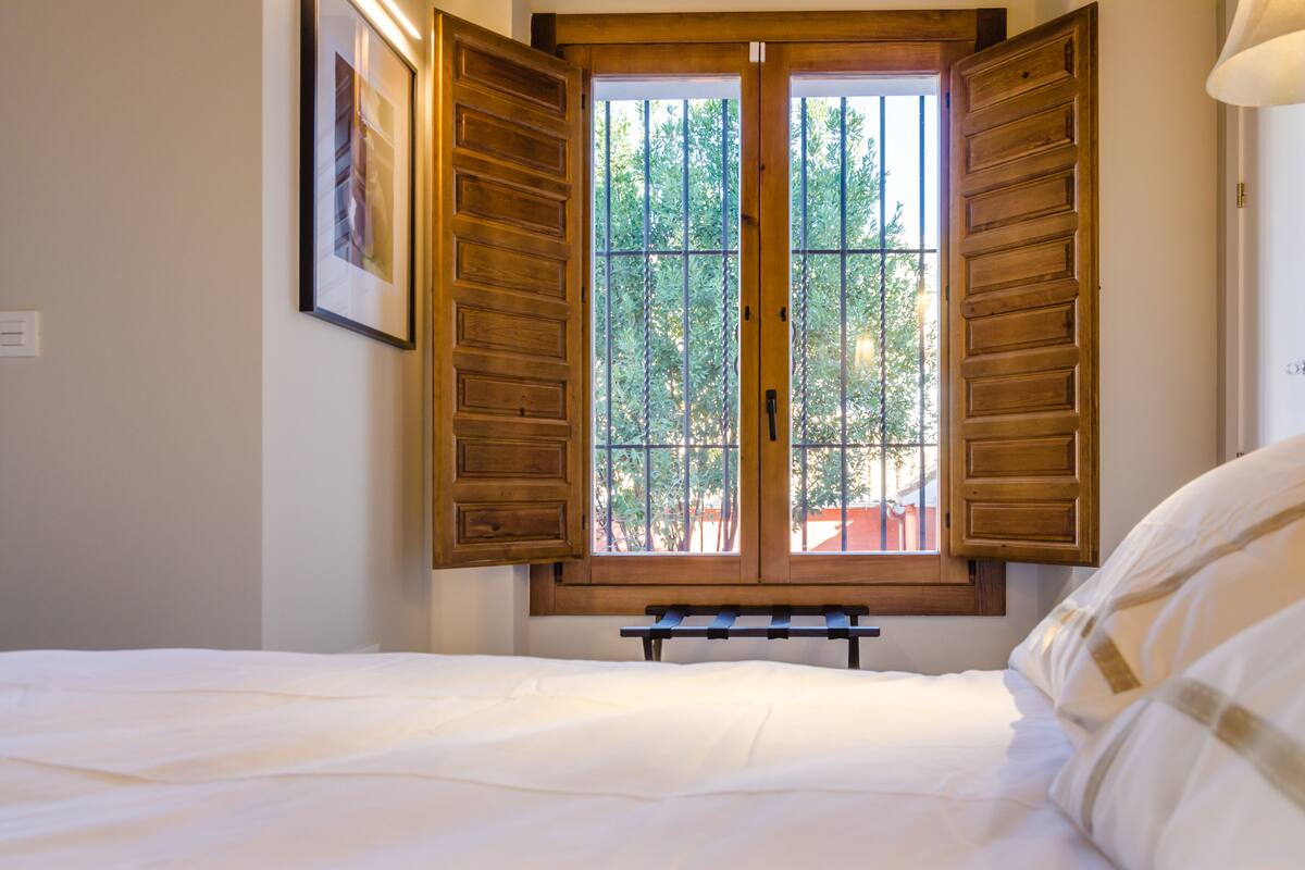 A bedroom with a bed, a wooden window with shutters, and a view of trees outside.