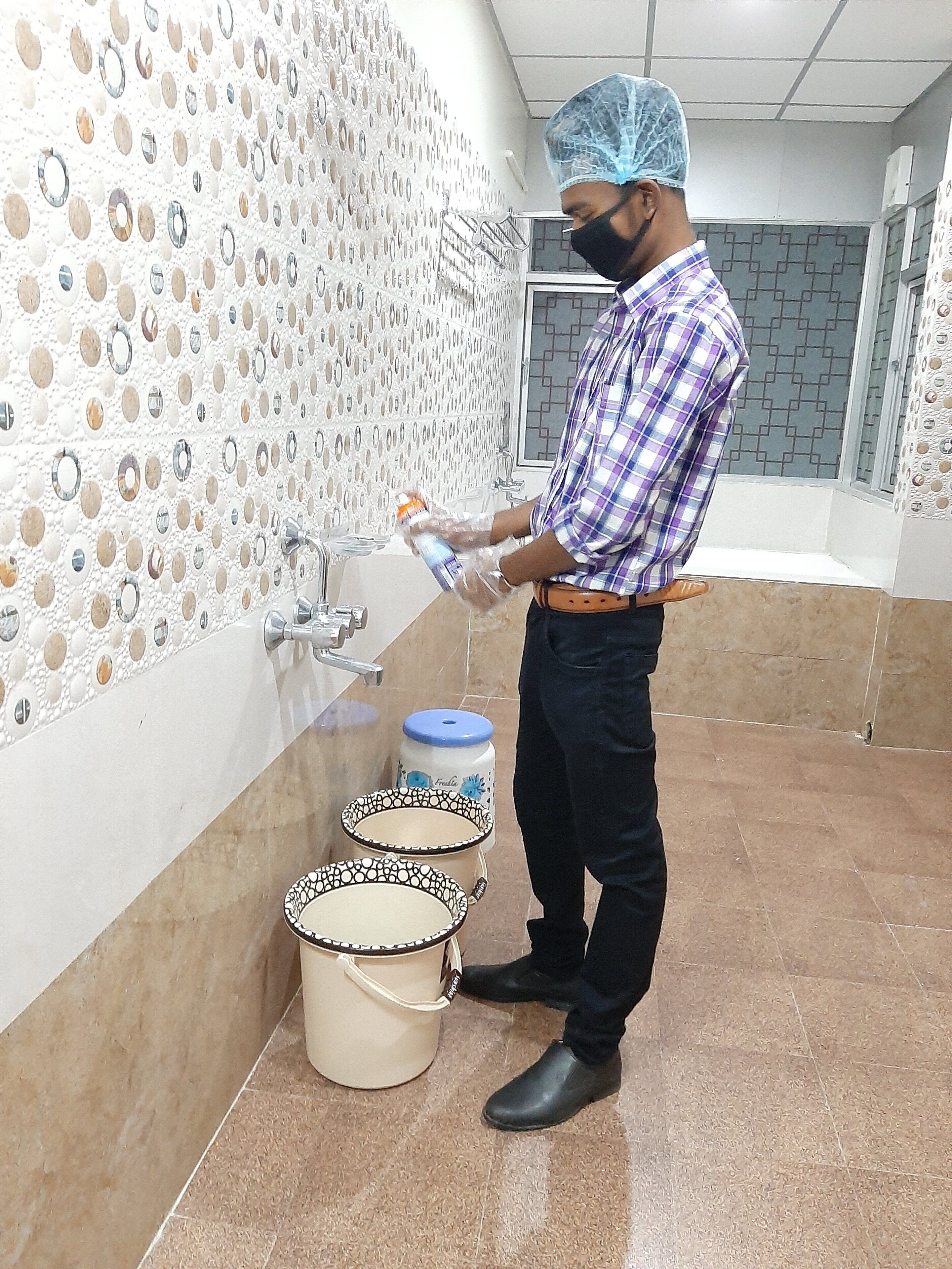 A person in a checkered shirt and black pants, wearing a hairnet, handling a bottle near a sink with a decorative wall. There are two large containers with lids on the floor.