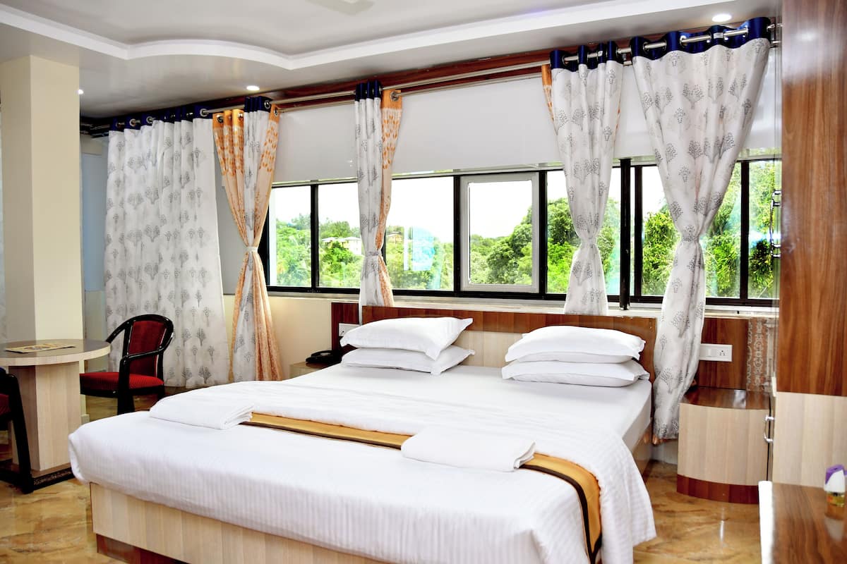 A hotel room with a large bed, a desk, and a chair. There are curtains on the windows, and the room has a view of greenery outside.