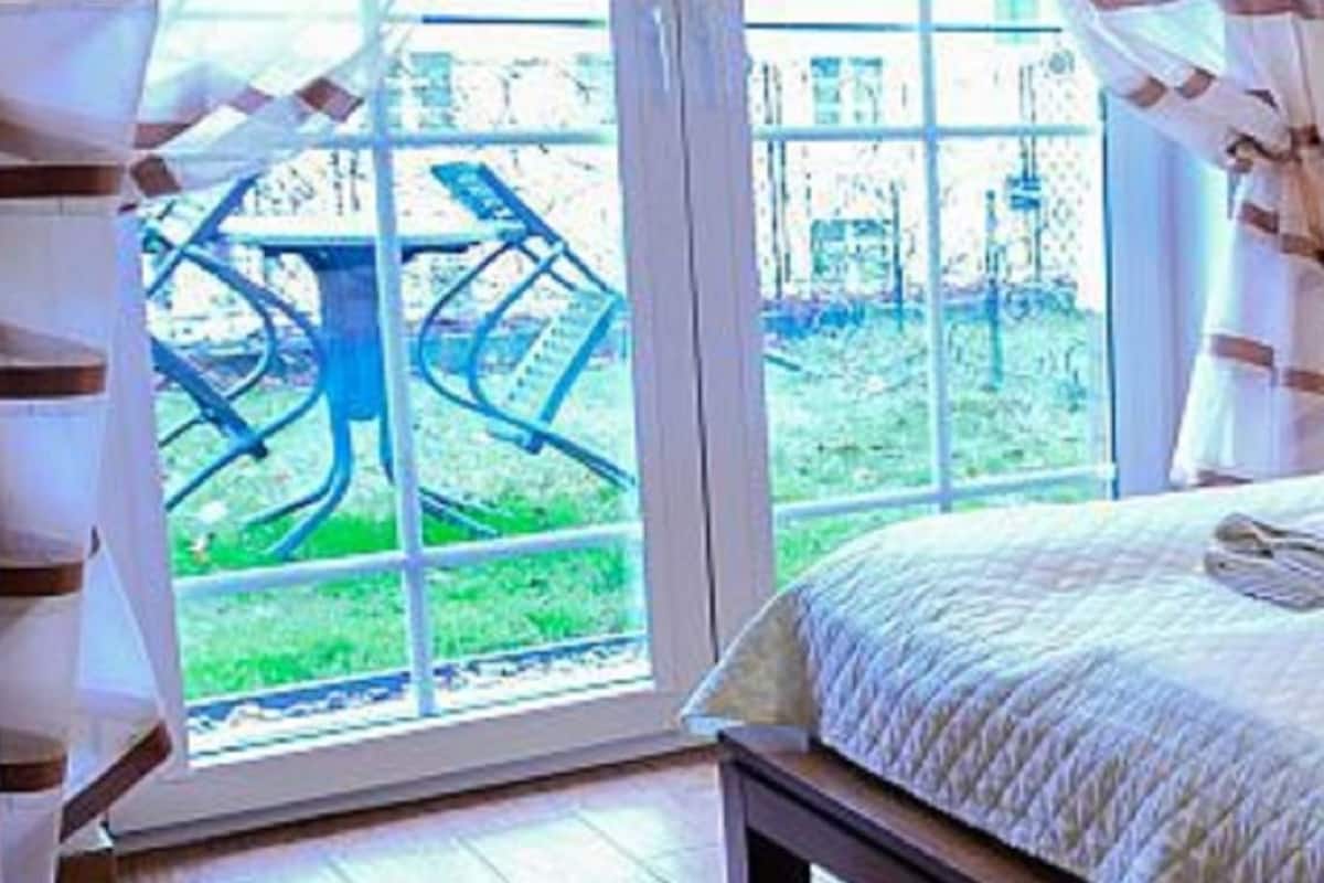 Quarto com cama, janela com cortinas e vista para um jardim e um prédio lá fora.