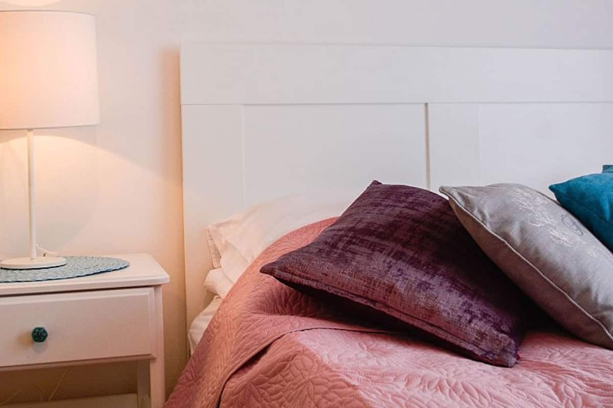Cama bem arrumada com colcha rosa, cabeceira branca e mesa de cabeceira com abajur.