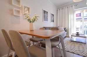 Uma sala de estar moderna com uma mesa de jantar de madeira, cadeiras estofadas cinzas, um sofá cinza e uma mesa de centro. Há obras de arte emolduradas na parede e um vaso com flores brancas sobre a mesa.