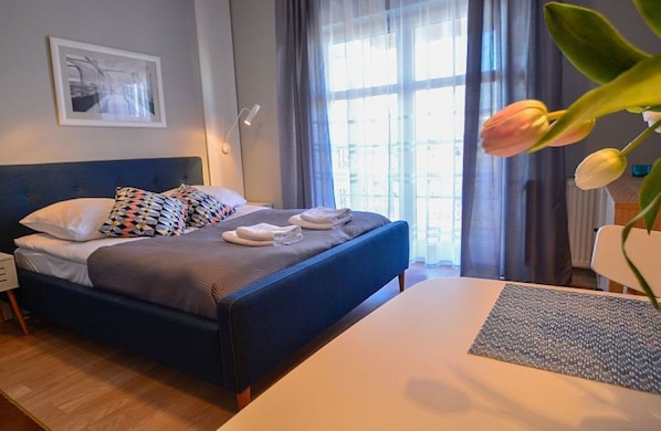 A neatly made bed with pillows, a bedside table, a lamp, and a window with curtains.
