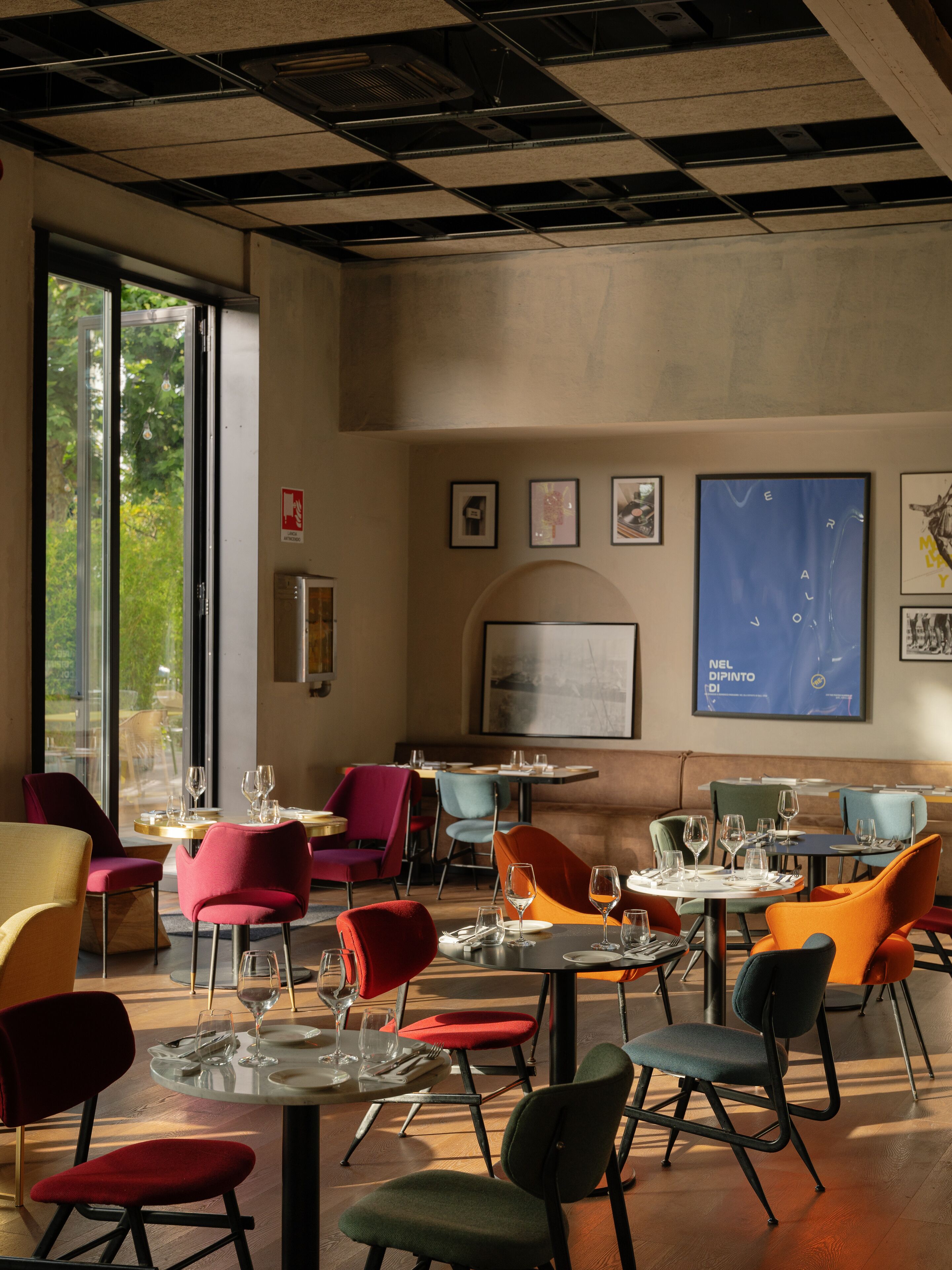 A modern dining area with round tables and colorful chairs, large windows, and artwork on the walls.