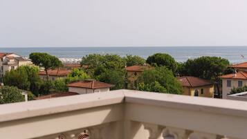 A coastal town with terracotta-roofed buildings, a clear view of the sea, and a balcony railing in the foreground.