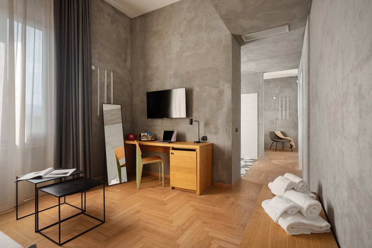 Une chambre d’hôtel moderne avec un bureau en bois, une chaise, un téléviseur à écran plat et une petite table. Des serviettes sont posées sur un banc, et la fenêtre donne sur un couloir.