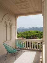 Un balcón con dos sillas verdes, barandilla blanca y vistas a las montañas.