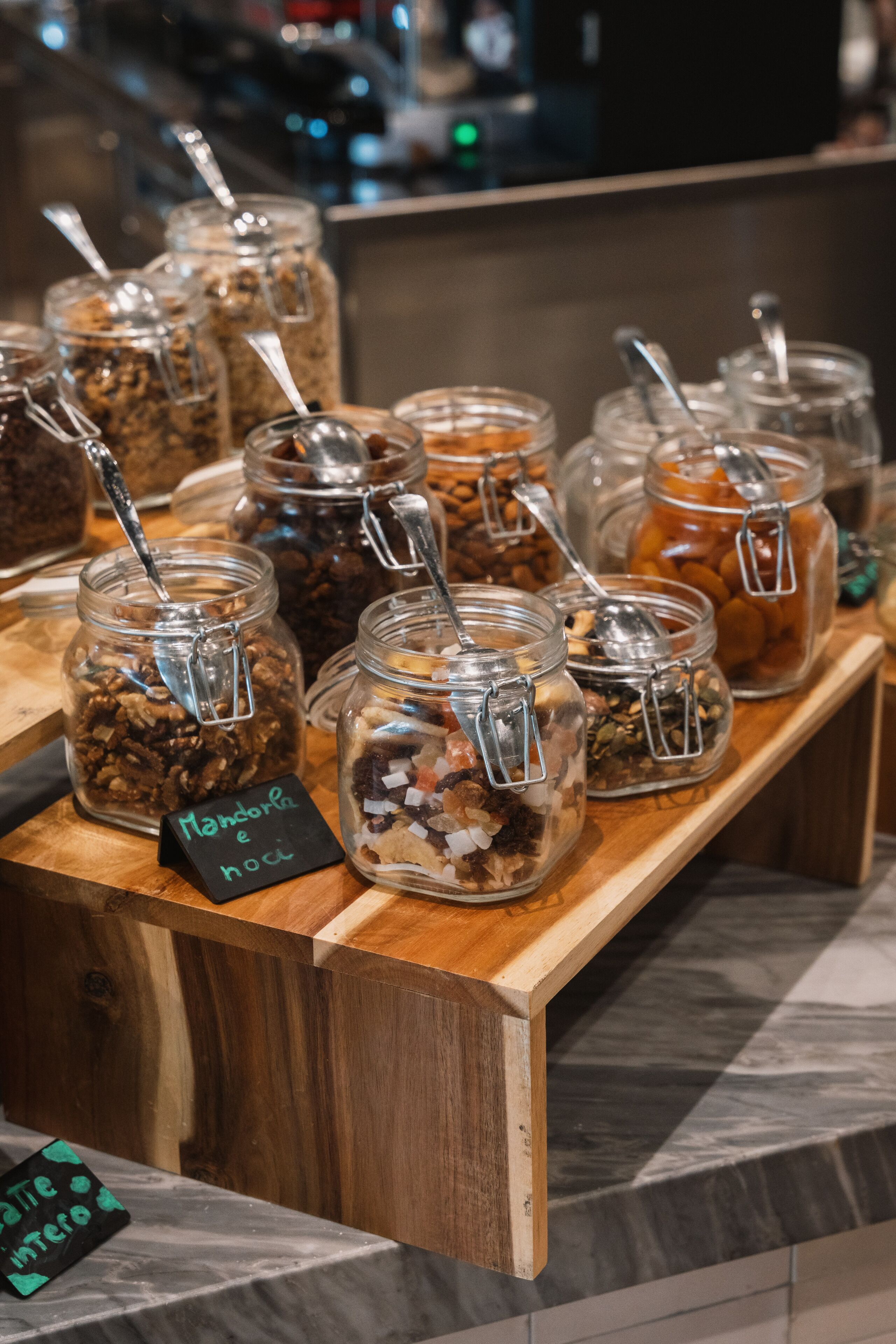 Una barra de madera con varios frascos de vidrio que contienen diferentes alimentos, cada uno con una cuchara de metal.